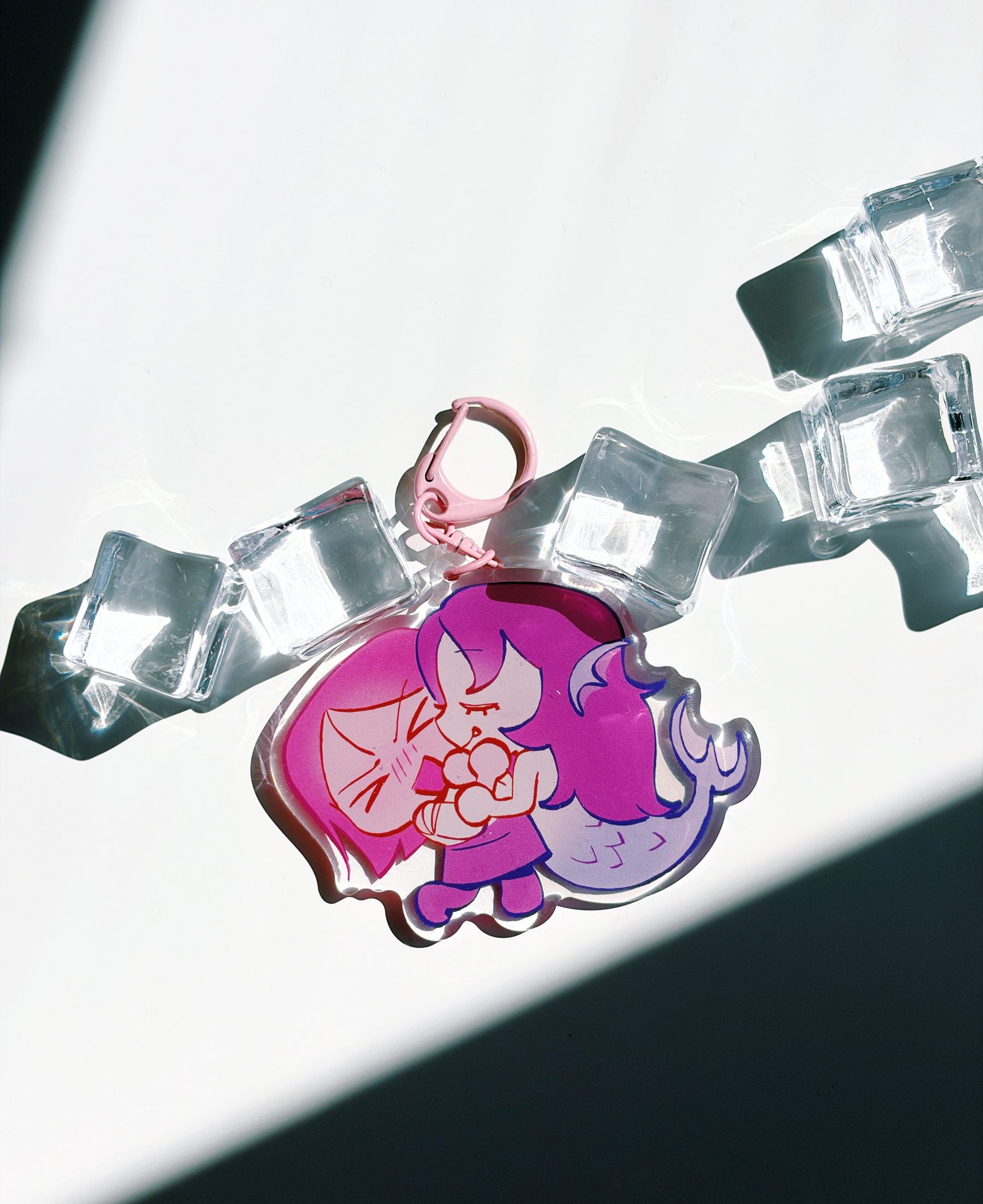 A pink and purple acrylic charm featuring an anime-style character holding a flower, attached to a pink clasp and photographed on a reflective chain with clear ice cubes against a bright white background with dramatic shadows.